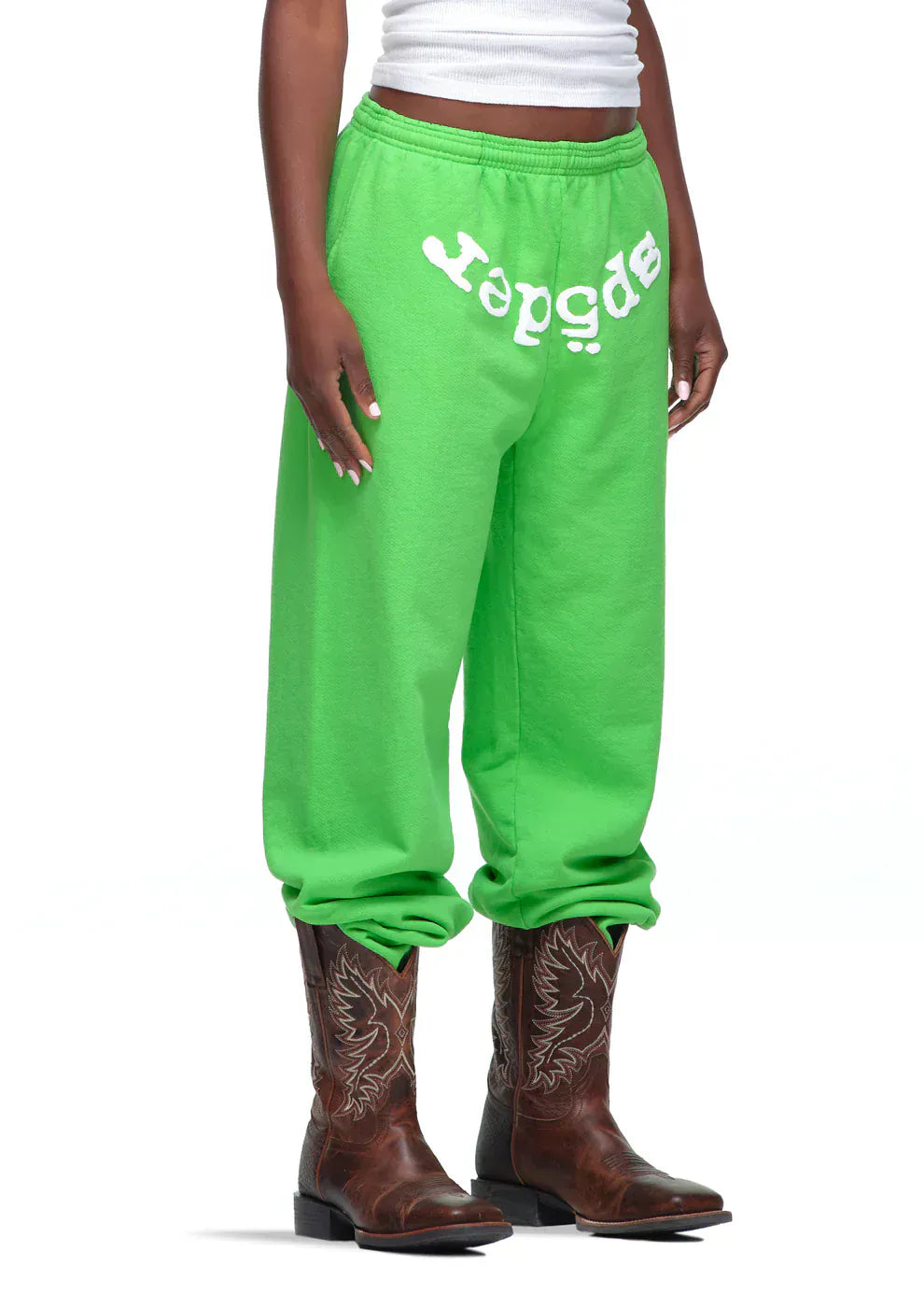 A person wearing Sp5der Sweatpants – Unisex Green Cotton Sweatpants with a front logo print, paired with a white tank top and brown cowboy boots.