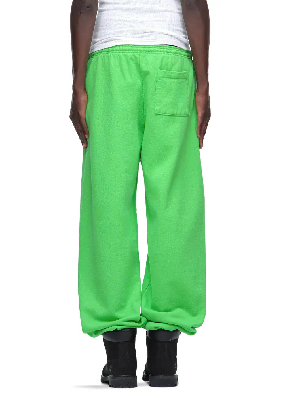 A person stands with their back to the camera wearing Sp5der Sweatpants—unisex green cotton sweatpants with a front logo print—paired with a white top and black shoes, highlighting a relaxed streetwear joggers style.