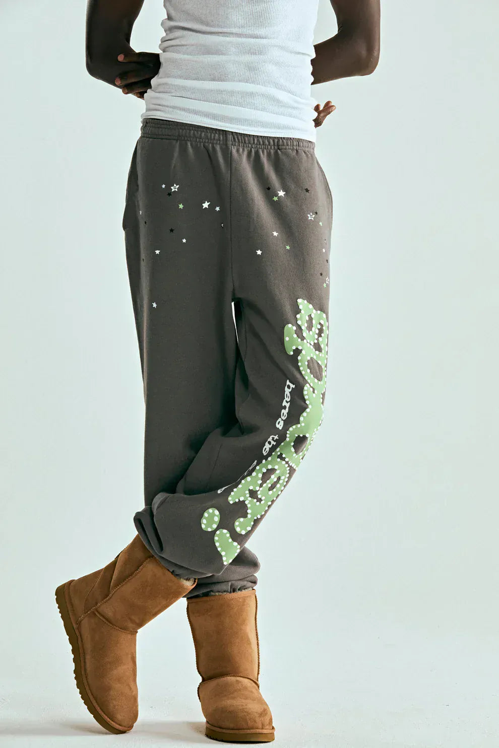 Person wearing Sp5der Sweatpants – Unisex Dark Grey Cotton Sweatpants with a green side logo and star print, paired with a white tank top and tan boots, standing against a plain background for a standout streetwear look.