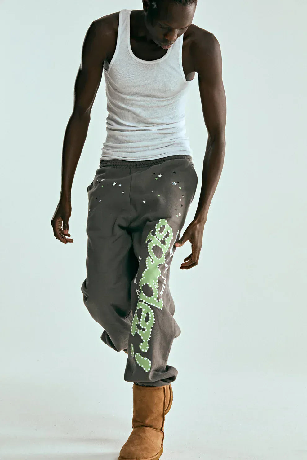 A person in a white tank top, tan boots, and Sp5der Sweatpants—unisex dark grey cotton pants with green side logo and star print—stands against a plain background, looking down and highlighting bold streetwear style.