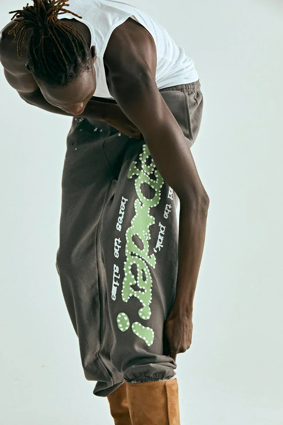Person in a white tank top wears Sp5der Unisex Dark Grey Cotton Sweatpants with green side logo and star print, paired with tan boots, as they bend to adjust the pant leg against a plain background, highlighting streetwear style.