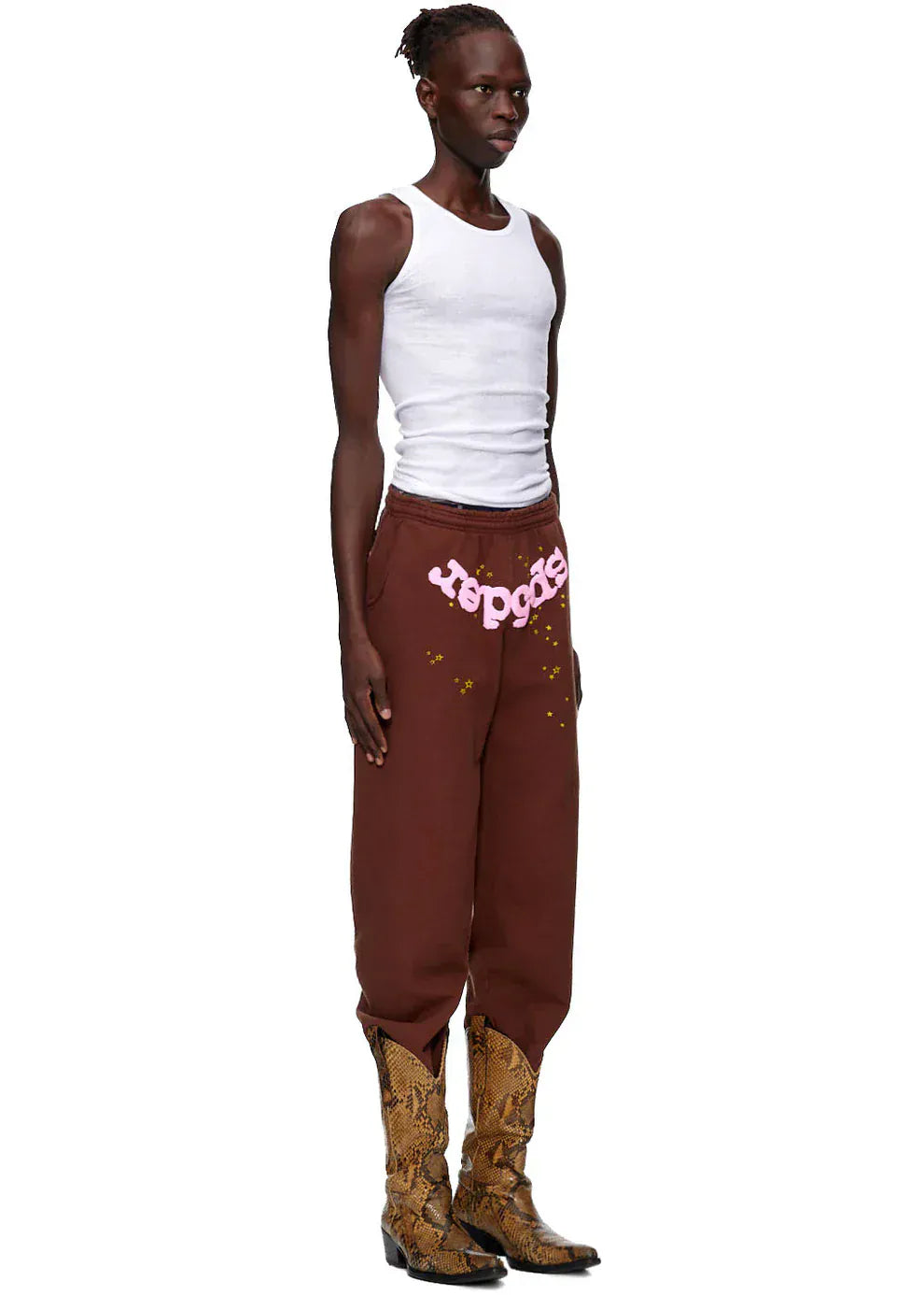 A person wearing a white tank top, Sp5der Sweatpants—unisex brown cotton sweatpants with a pink front logo and star print—and brown snakeskin boots stands against a plain background.