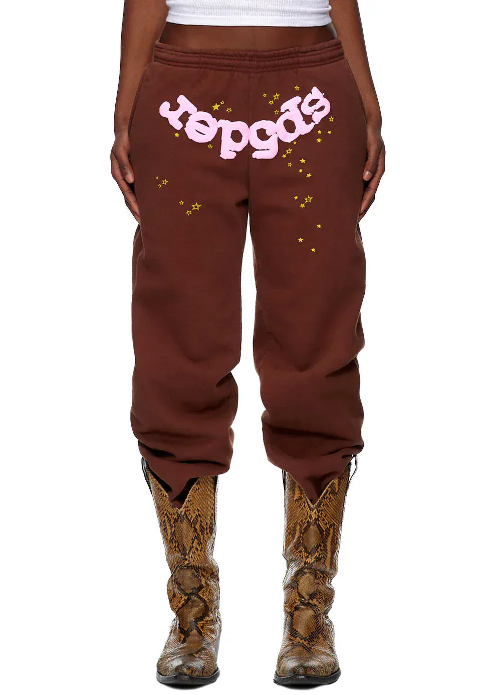 Someone wearing Sp5der Sweatpants—unisex brown cotton sweatpants with a pink front logo and star print—paired with brown snakeskin boots.
