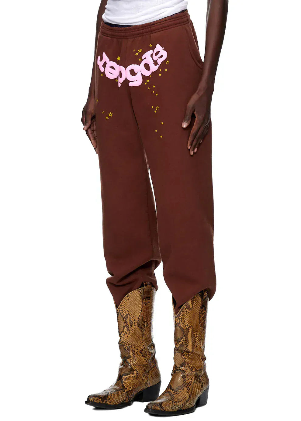 A person wearing Sp5der Sweatpants – Unisex Brown Cotton Sweatpants with a pink front logo and star print, paired with a white shirt and brown snakeskin cowboy boots.