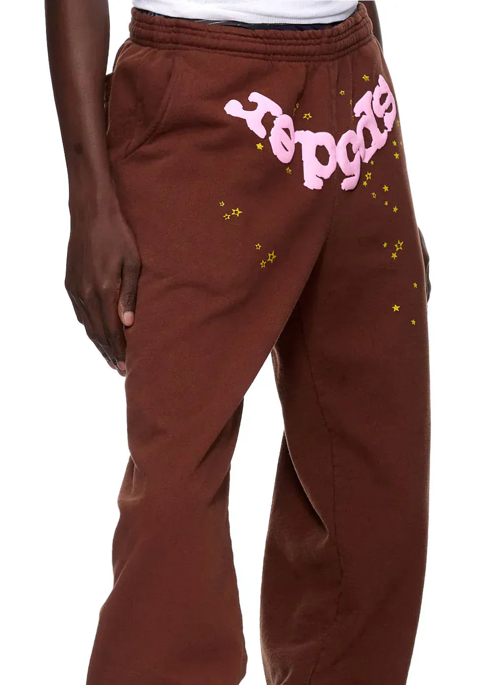 A person wears Sp5der Sweatpants—unisex brown cotton with a pink front logo and yellow star print—shown from waist to mid-calf.