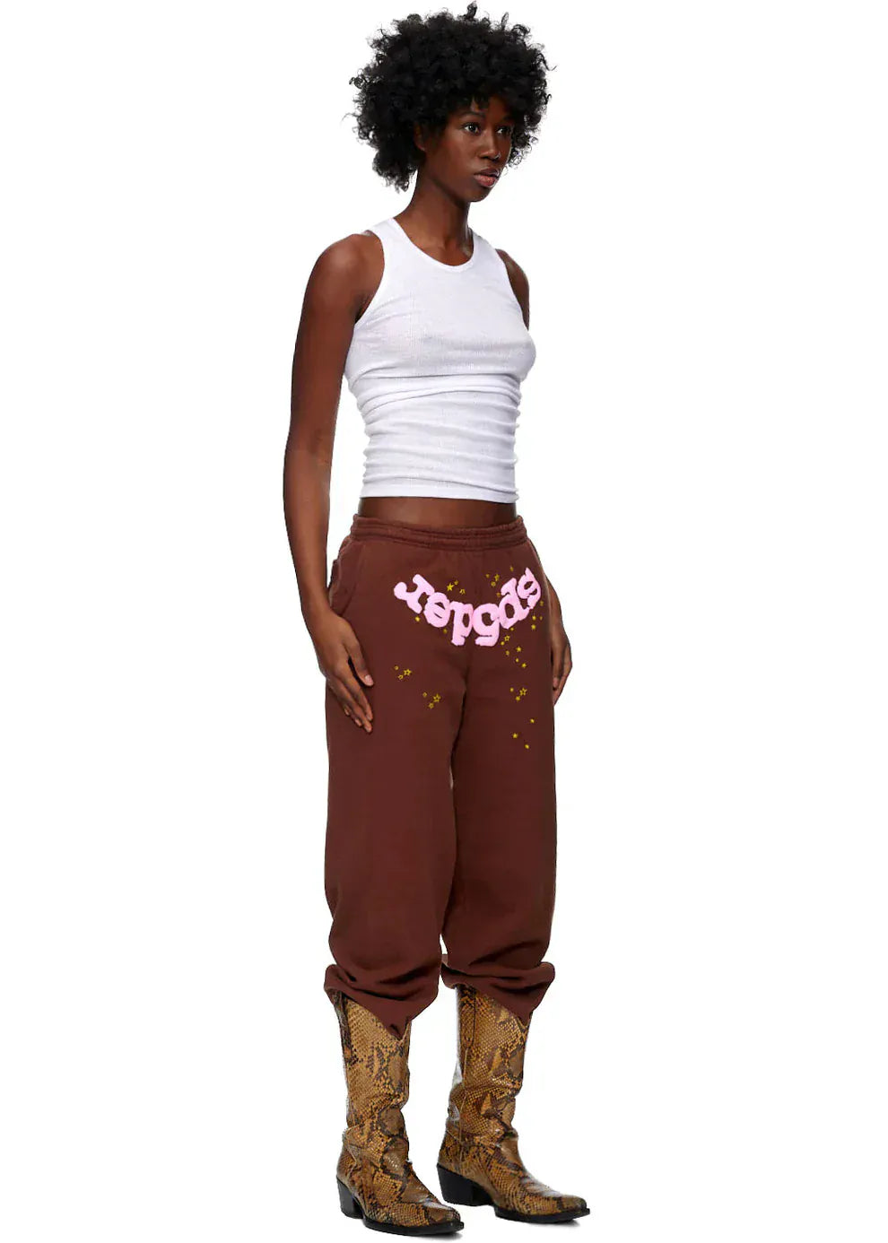 A woman stands slightly turned, wearing a white tank top, Sp5der Sweatpants—unisex brown cotton joggers with a pink front logo and star print—and snake print cowboy boots.