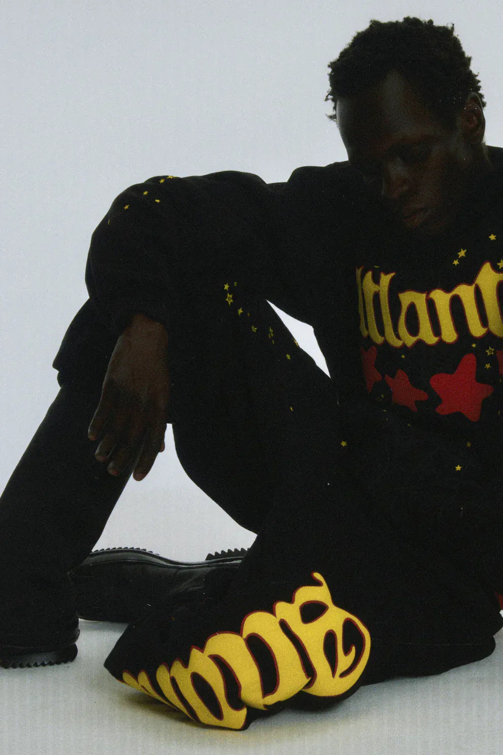 A person sits on the floor wearing Sp5der Sweatpants—unisex black cotton sweatpants featuring a flame side logo and star print—paired with a graphic top, set against a plain light background.