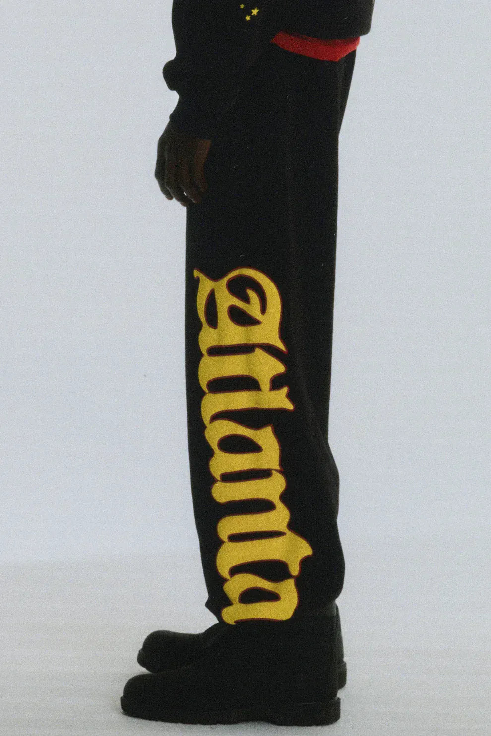 A person wearing Sp5der Sweatpants – unisex black cotton joggers with a flame side logo and star print, paired with black shoes for an effortlessly cool streetwear look.