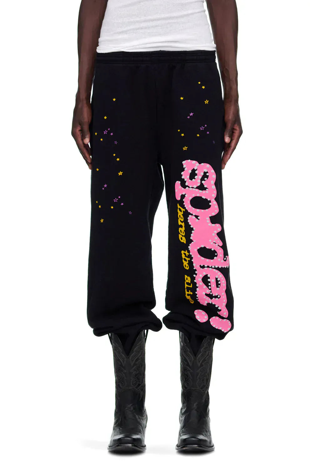 Wearing Sp5der Sweatpants – unisex black cotton sweatpants with pink graphic logo and star print – with black cowboy boots and a white top creates a bold streetwear look.