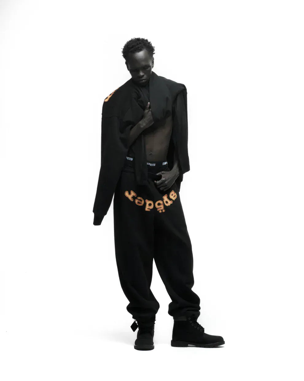 A person stands against a white background wearing black Sp5der Sweatpants – unisex black cotton sweatpants with a flame front logo print – and a matching black jacket draped over their shoulders.