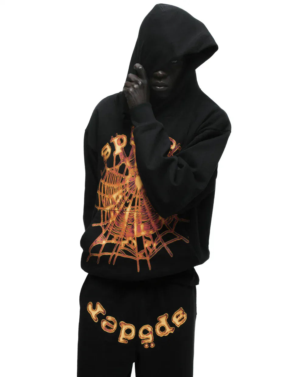 A person in a black hoodie and Sp5der Sweatpants – Unisex Black Cotton Sweatpants with Flame Front Logo Print stands against a white background, their face partly hidden by the hood.