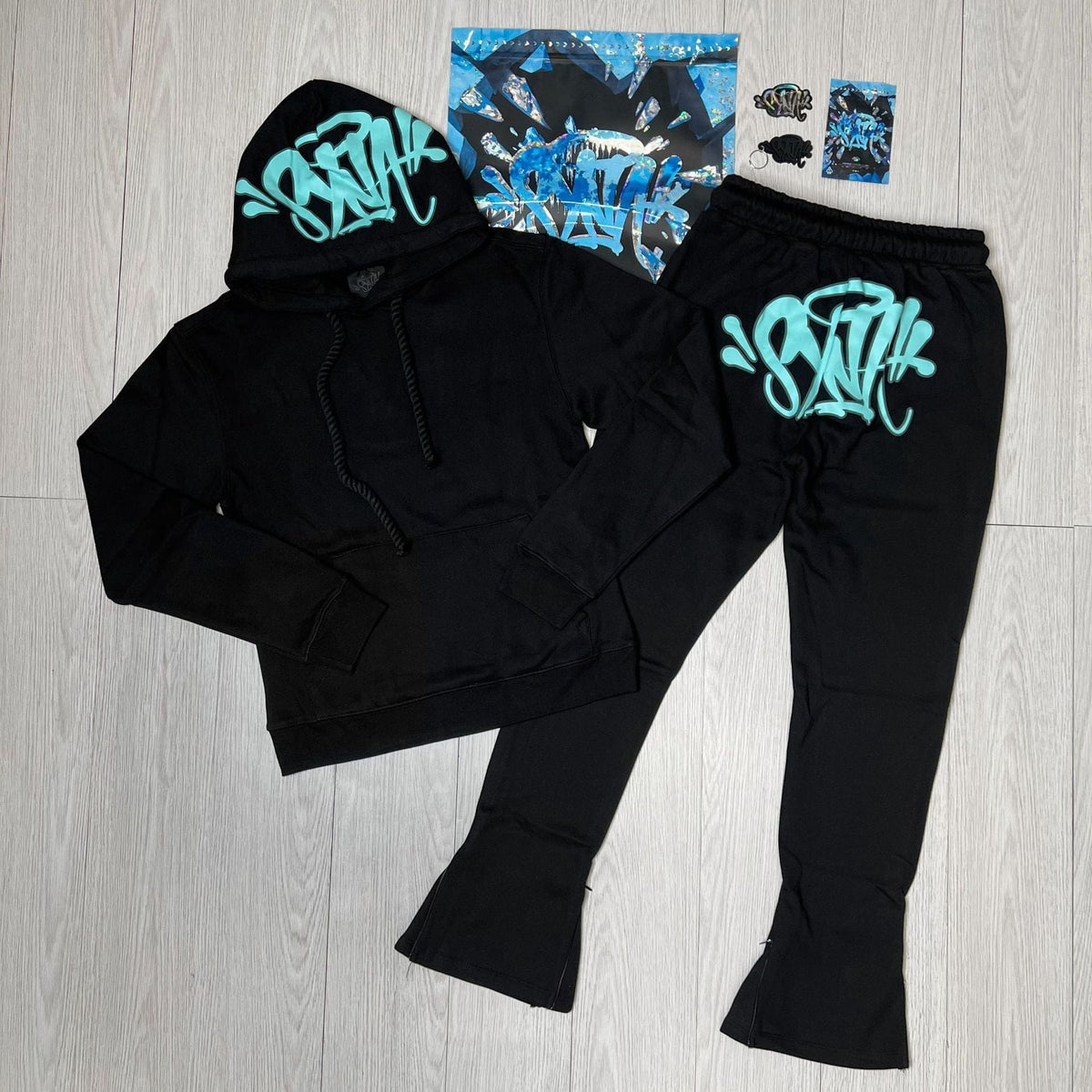 Syna World Graphic Hoodie and Flared Pants Tracksuit Set with Accessories, shown in black with turquoise graffiti-style text, laid out on a light wood floor alongside matching stickers and printed streetwear items. Available in 4 colors.