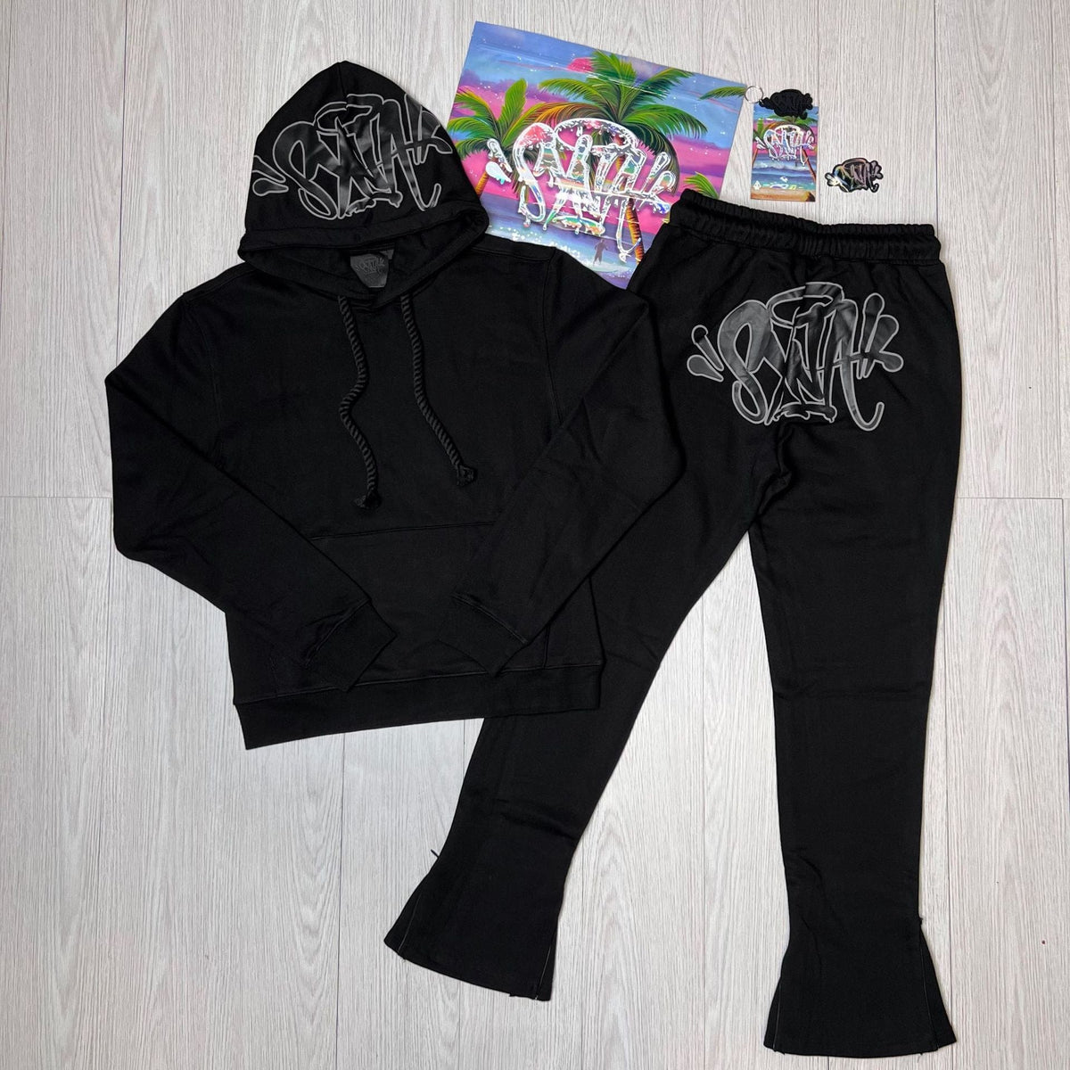 Syna World Tracksuit Set—Graphic Hoodie & Flared Pants with Accessories—featuring white graffiti text, displayed on a light wood floor next to colorful items with a tropical print.