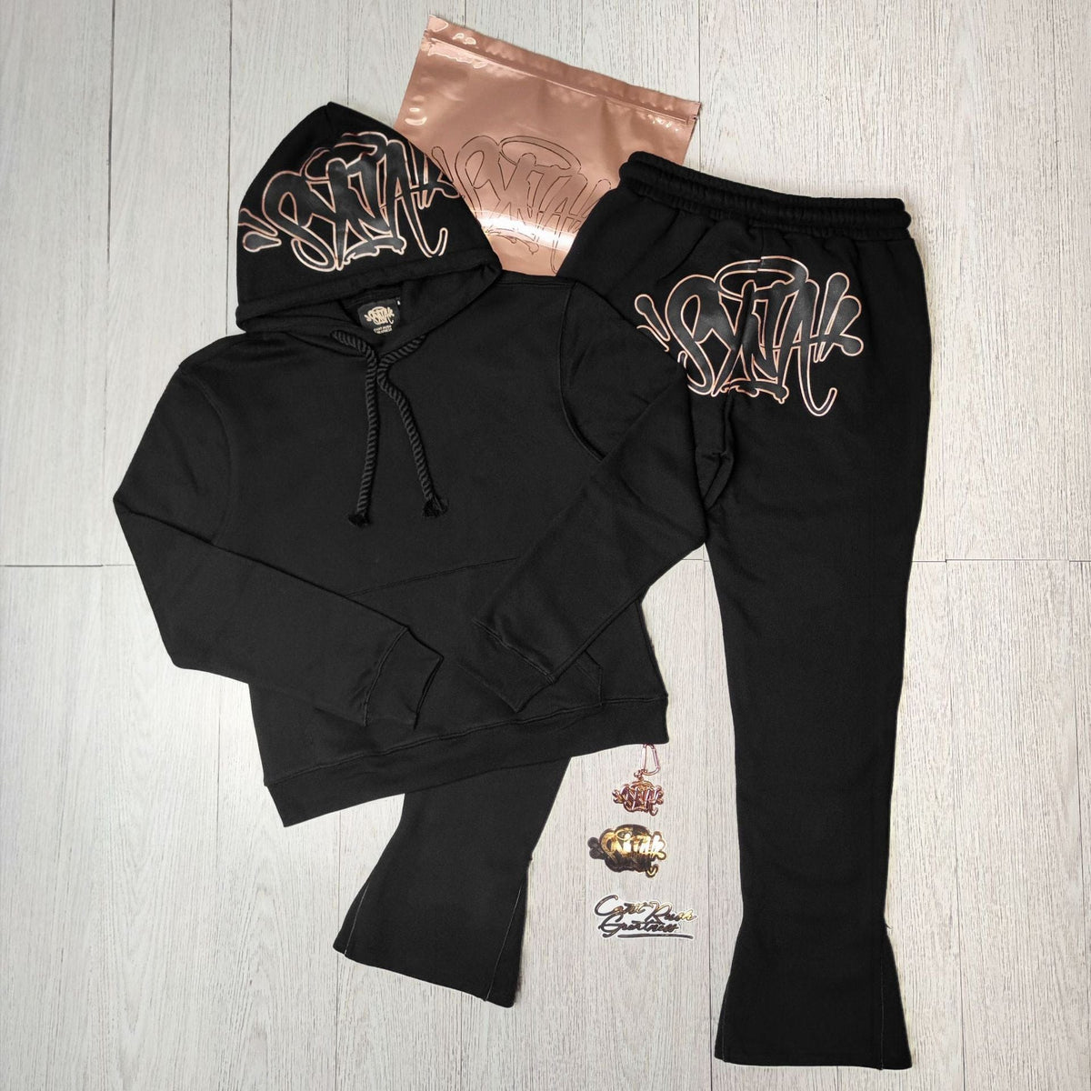 The Syna World Graphic Hoodie and Flared Pants Tracksuit Set, shown in black with a bold graffiti logo, is displayed on a light floor with accessories including a metallic bag, stickers, and pins. Available in four colors.