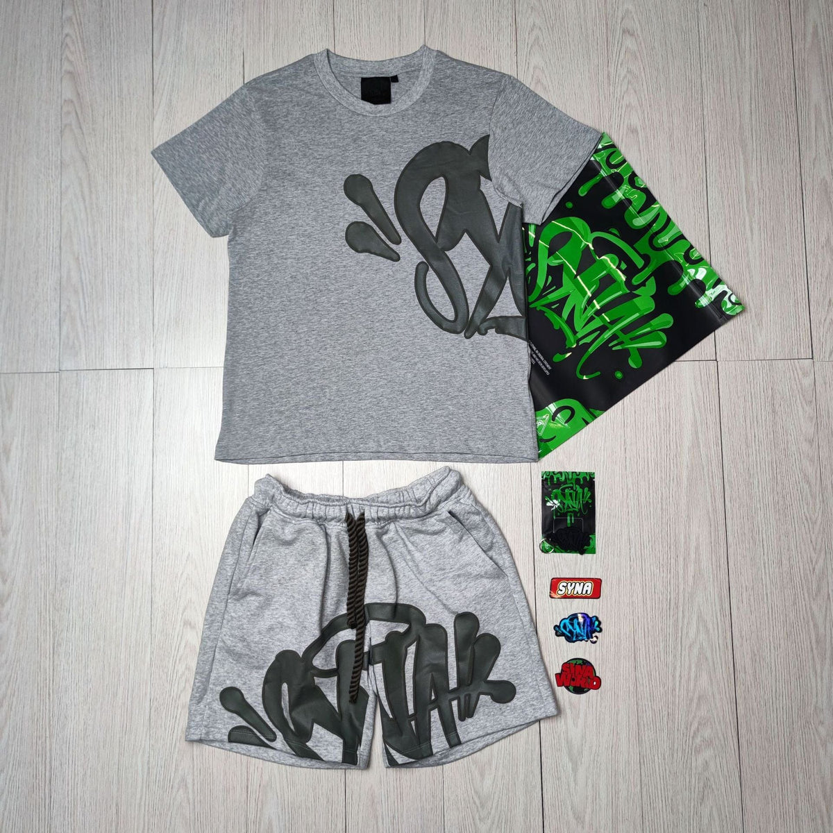 The Syna World T-Shirt and Shorts Set with Accessories – 8 Colors features gray pieces with black and green graffiti-style lettering, a matching green and black bag, plus four assorted streetwear pins or patches.