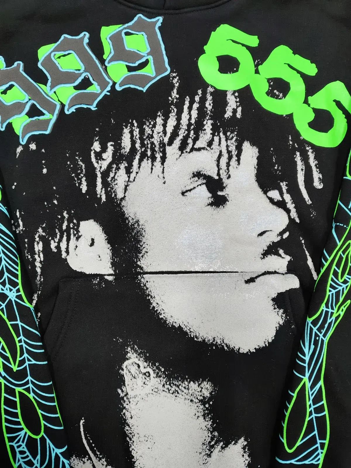 The Sp5der “999 555 Legacy” Hoodie in black features a grayscale portrait, "999" in blue, "666" in green, blue-green web sleeve details, and is made from 440GSM heavyweight cotton fleece for comfort. Inspired by the classic Sp5der style.