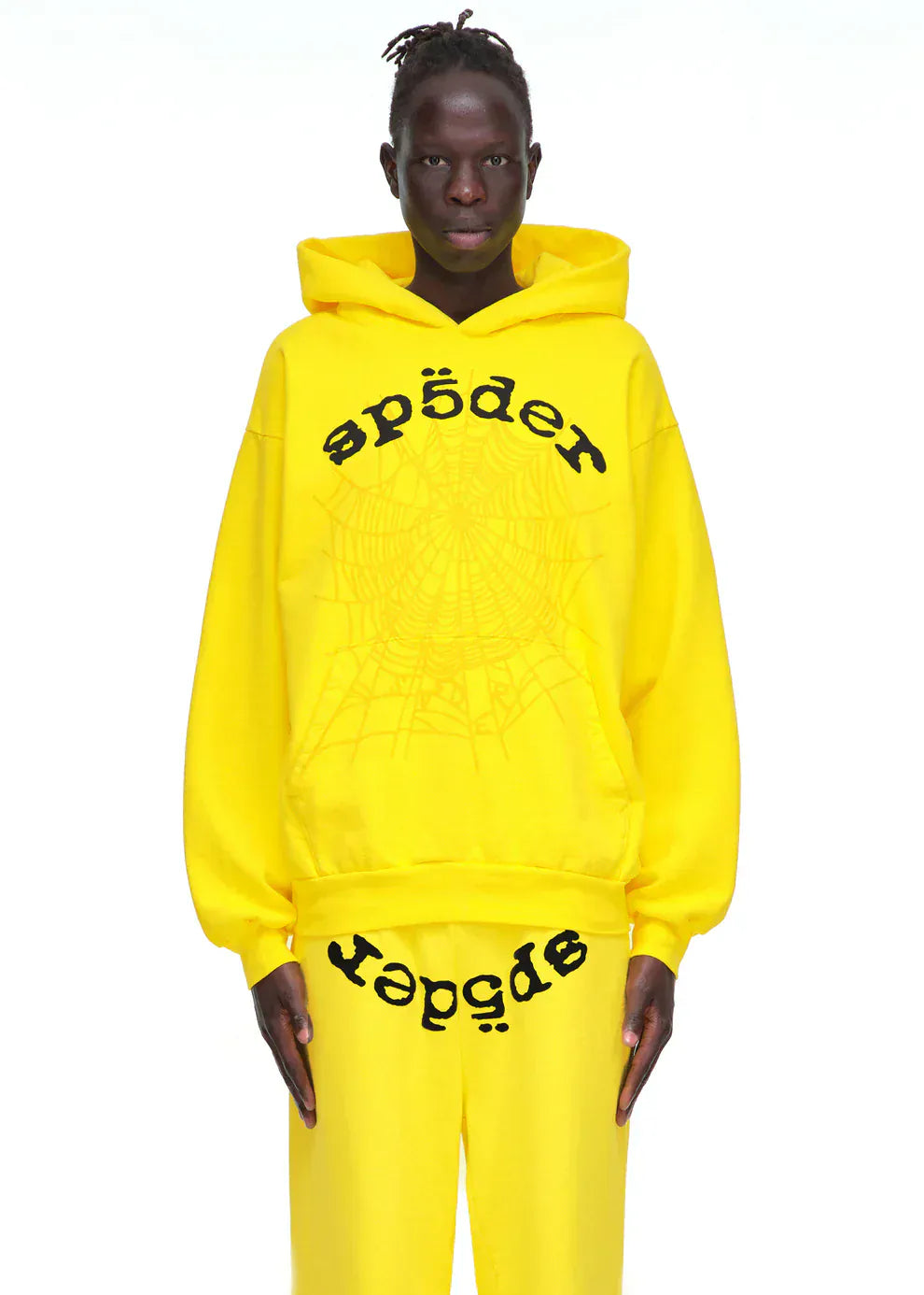 A person models the Yellow Graphic Hoodie with Spider Web Print and Lettering – Comfort Fit Pullover Hoodie and matching pants, styled as bold streetwear, against a plain white background.