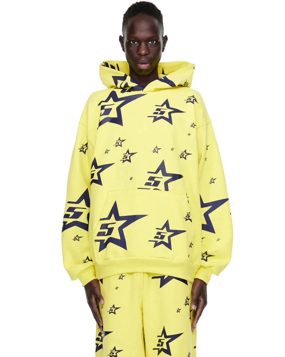 A person wears the Yellow All-Over Star Graphic Hoodie – Comfort Fit Pullover Hoodie and matching pants, featuring large and small black star designs, while standing against a plain background.