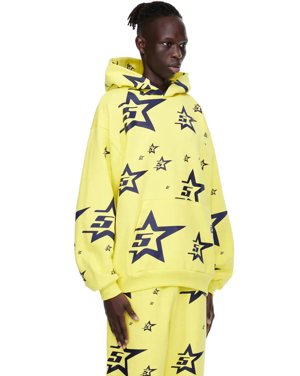 A person wears the Yellow All-Over Star Graphic Hoodie – Comfort Fit Pullover Hoodie with matching star-patterned pants, standing against a plain white background.