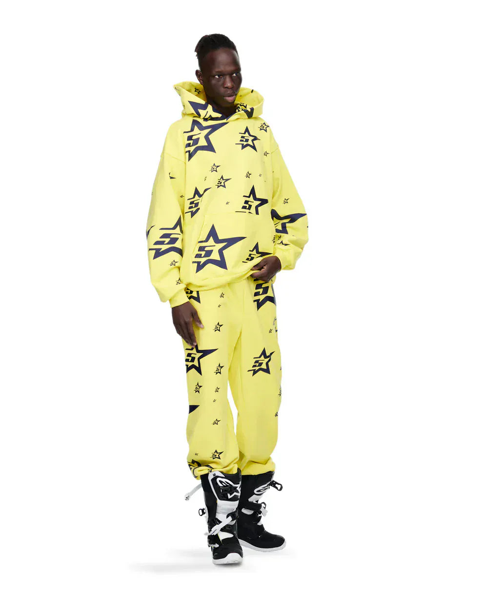 A person wears the Yellow All-Over Star Graphic Hoodie – Comfort Fit Pullover Hoodie with matching pants and black and white boots, standing against a plain white background.