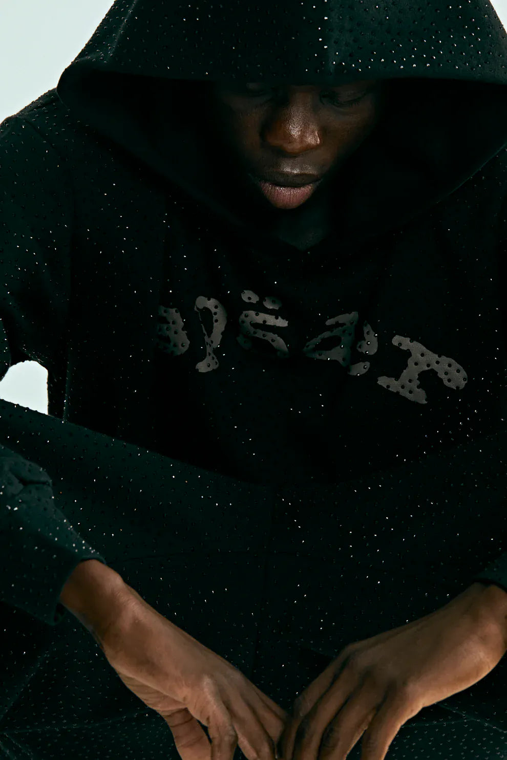 A person wearing a VVS Spider Hoodie – Black Sp5der Rhinestone Pullover with Diamond Graphic Detail, head slightly down and hands resting in front.