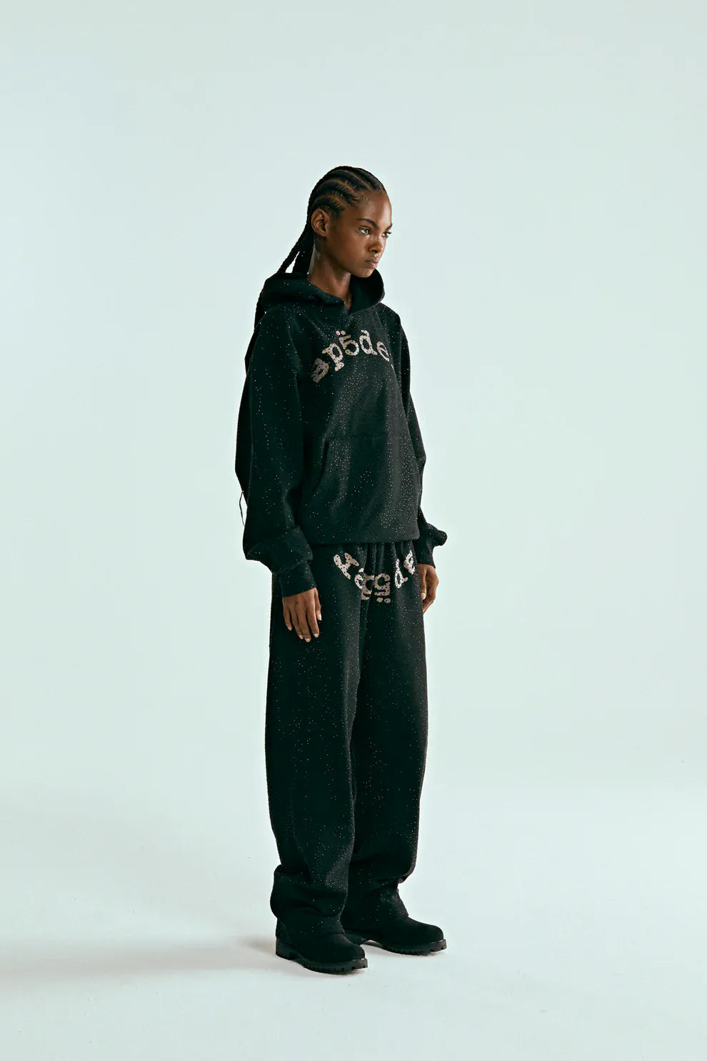 A person wears the VVS Spider Hoodie – Black Sp5der Rhinestone Pullover with diamond graphic detail, paired with matching sweatpants and black shoes, standing against a plain light background.