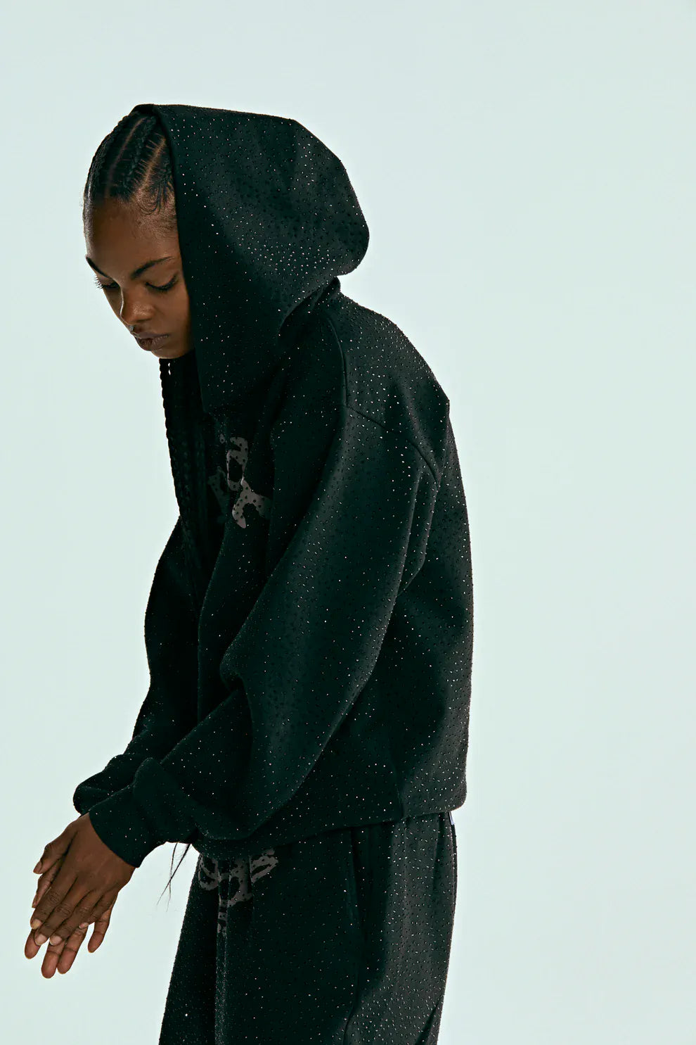 Person wearing the VVS Spider Hoodie – Black Sp5der Rhinestone Pullover with Diamond Graphic Detail and matching pants featuring small white speckles, looking downward against a plain light background.