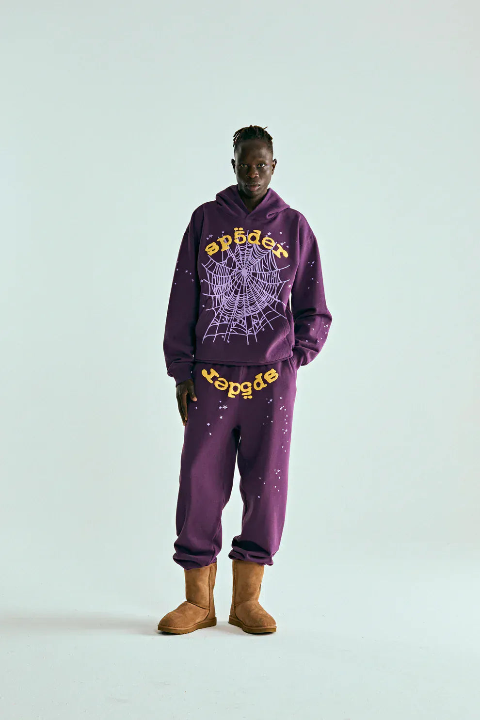 A person stands against a plain background wearing the Sp5der Web Hoodie – Deep Purple & Yellow 440GSM Heavyweight Oversized Streetwear Pullover and sweatpants with yellow web graphics, embracing luxury streetwear vibes, paired with tan slippers.