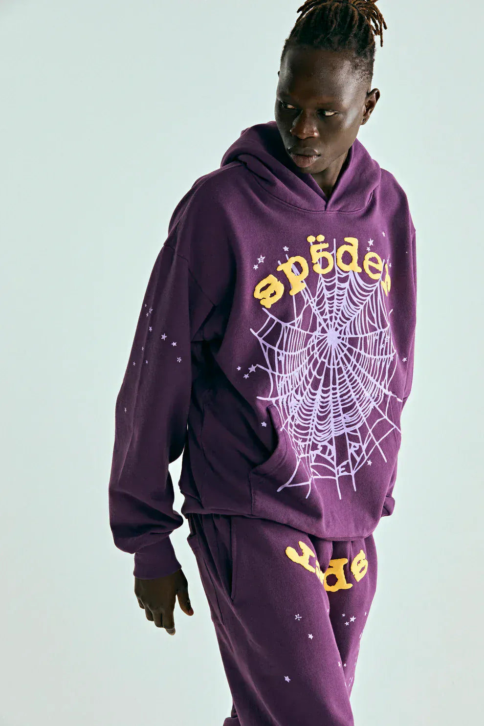 Person wearing the Sp5der Web Hoodie – Deep Purple & Yellow 440GSM Heavyweight Oversized Streetwear Pullover with matching pants, featuring a white spiderweb and yellow "sp5der" graphic, embodying luxury streetwear against a plain backdrop.